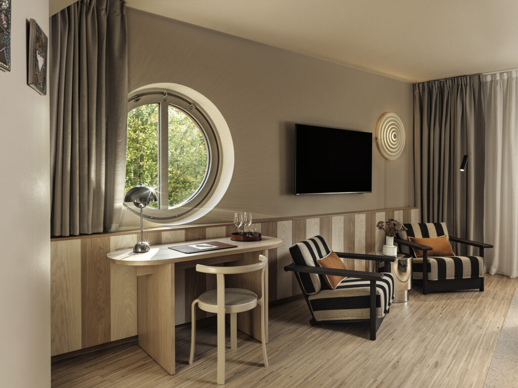 Ett sparsamt inrett hotellrum med tv, fotöljer och skrivbord, Ett runt fönster i väggen avslöjar en grönskande skog.
