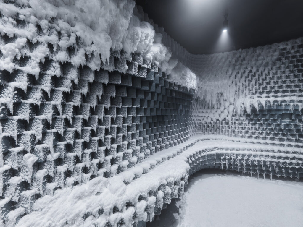 Ett mörkt rum med mönstrade väggar täckta av snö och frost. En lång bänk följer väggarna. 