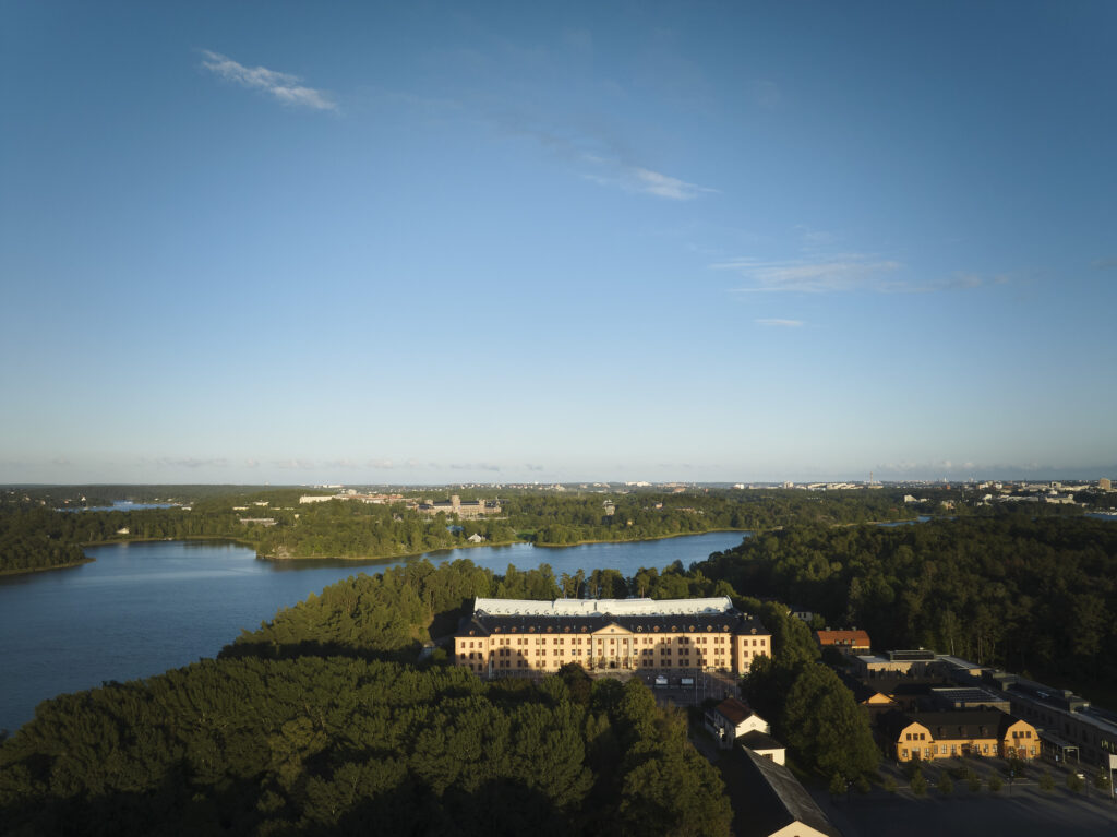 En stor hotellbyggnad i en grön skog, blått vatten syns under den blå himlen.