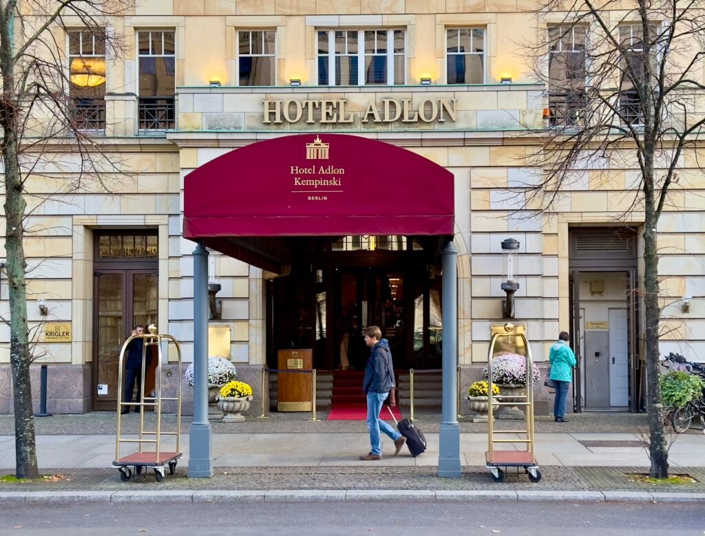 En röd markis hänger ovanför en entrétrappa till en hotellbyggnad. En man med rullväska går förbi utanför.