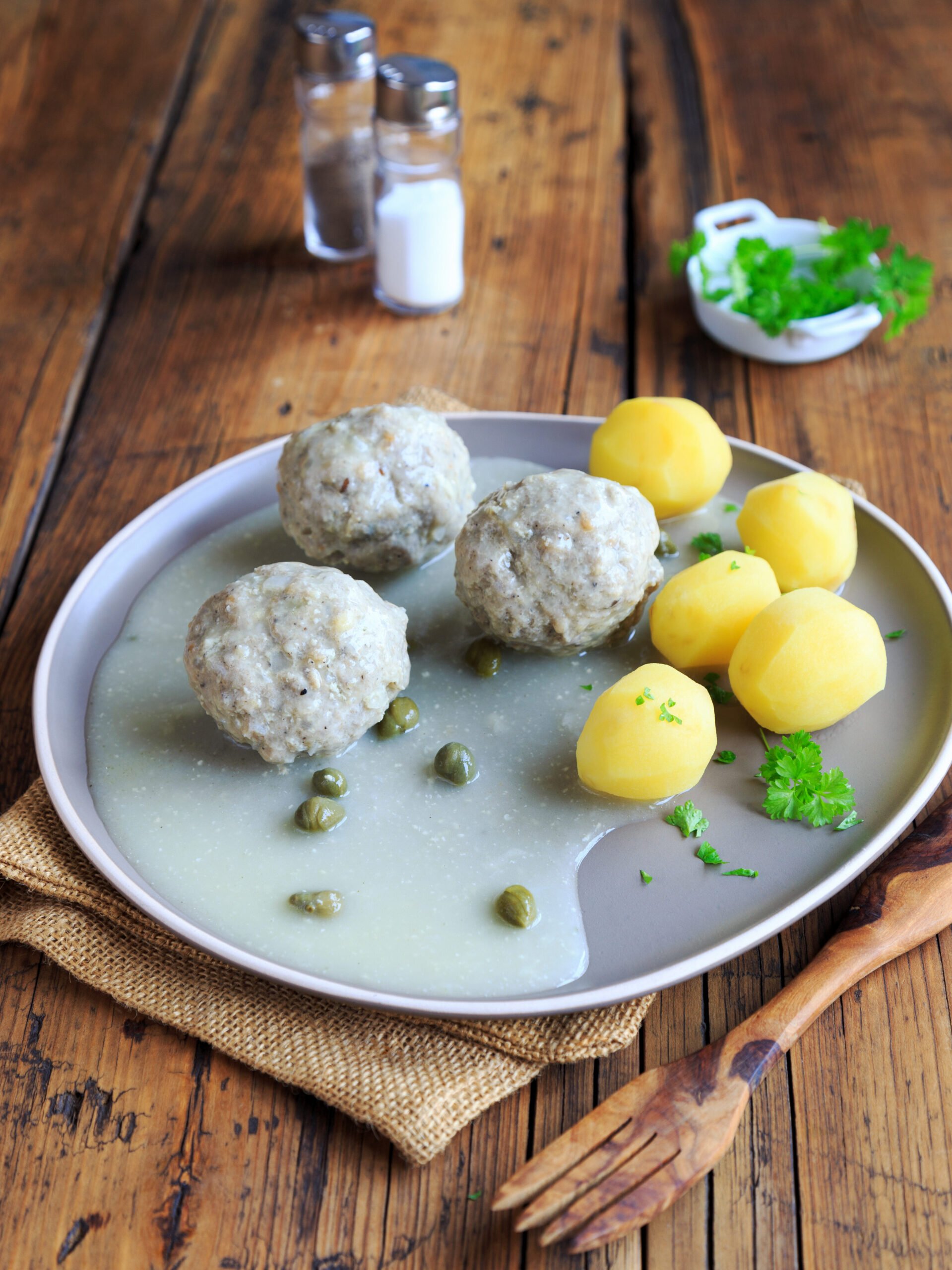 Bleka köttbullar ligger i en vit sås med kapris, på en tallrik tillsammans med kokta, skalade potatisar.
