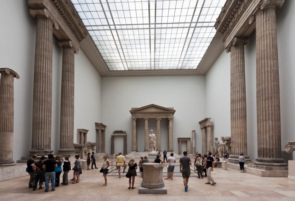 En stor museilokal med klassiska kolonner och högt i tak. Människor kikar på utställningen. 