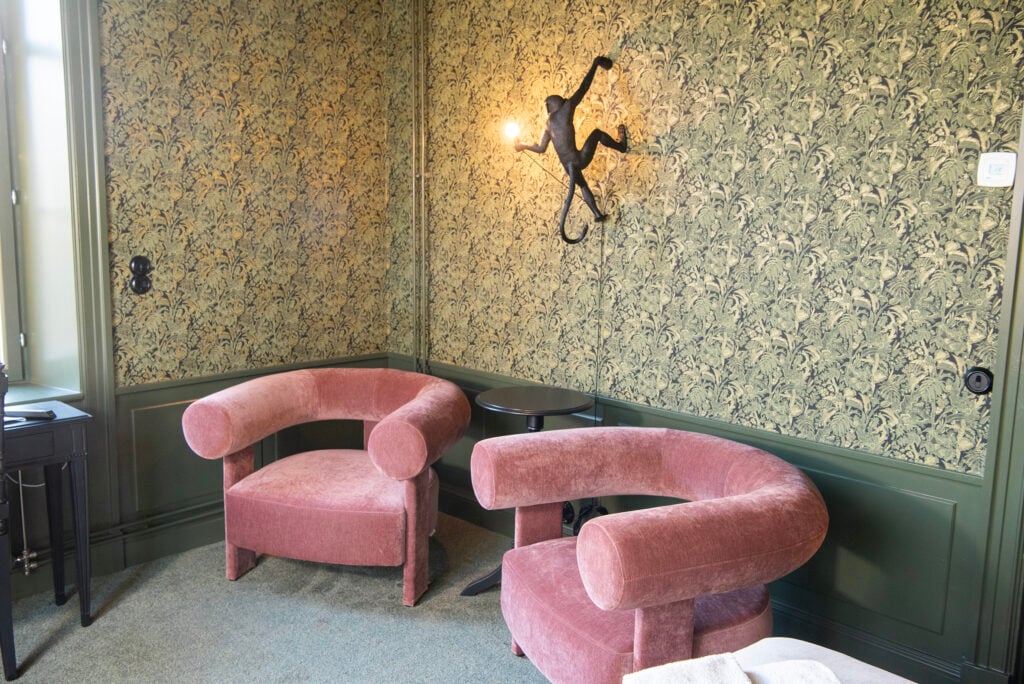 Två rosa plyschfåtöljer står mot en grön mönstrad vägg, en apformad lampa sitter på väggen ovanför. 