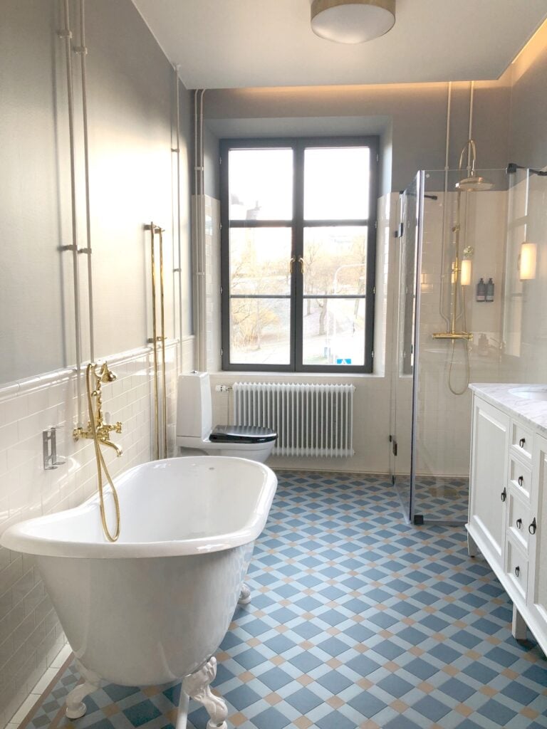 Ett vitt badkar med lejontassar och guldfärgade kranar, på ett blårutigt golv, en dusch i glas, ett fönster visar utsikten. 