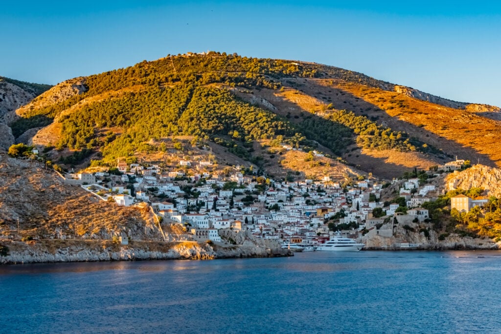 En grön kulle i gyllene solnedgångsljus, nedanför en by full av vita hus, havet framför.