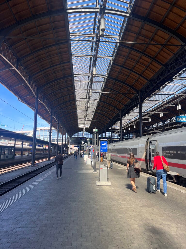 Människor går längs med en grå tågperrong, högt i tak, ett grått tåg med en röd rand står inne.