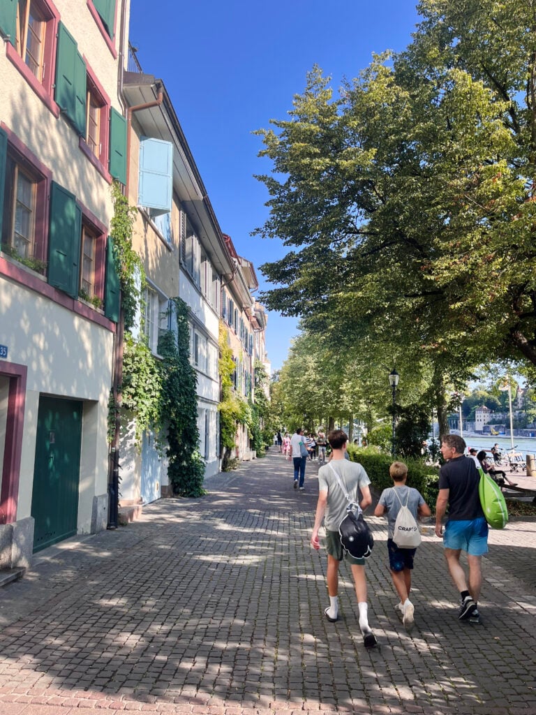En vuxen och två stora barn promenerar på en trottoar under gröna träd. 