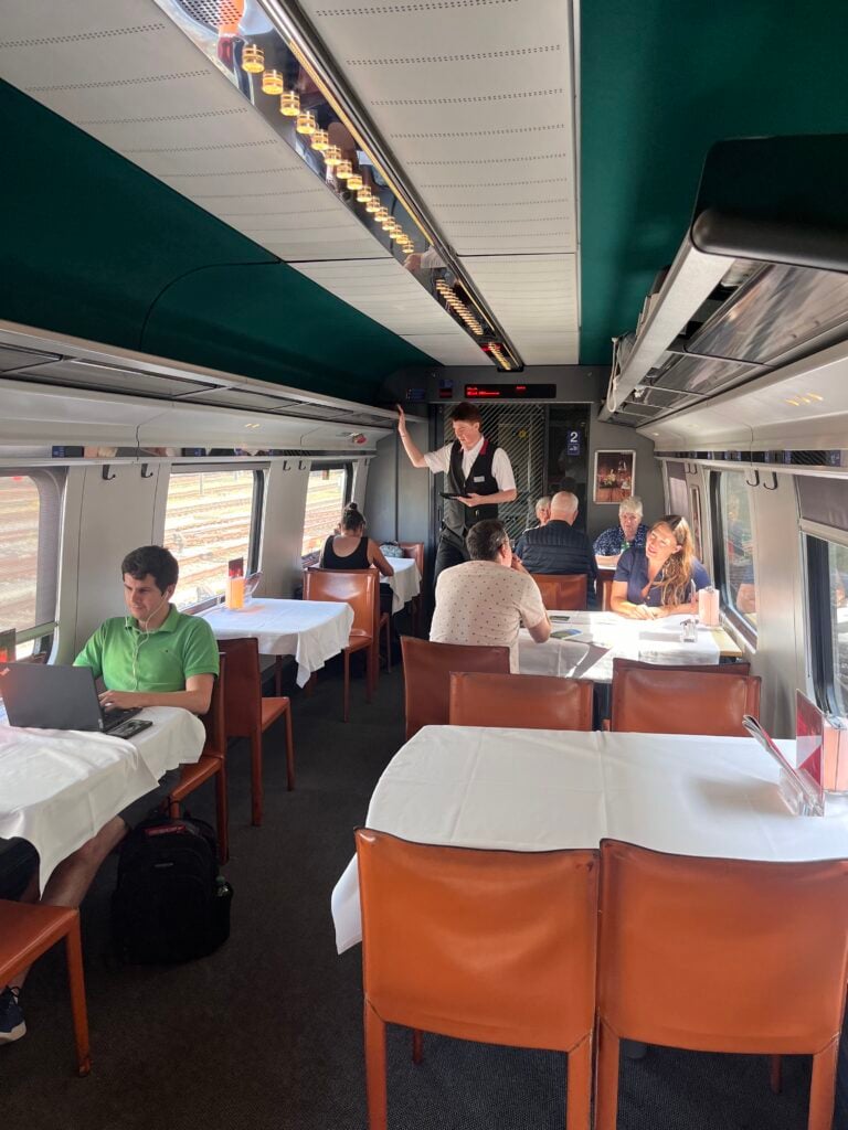 En tågvagn fylld med bruna skinnstolar och bord med vita dukar, Människor sitter och jobbar vid borden, en konduktör i gången. 