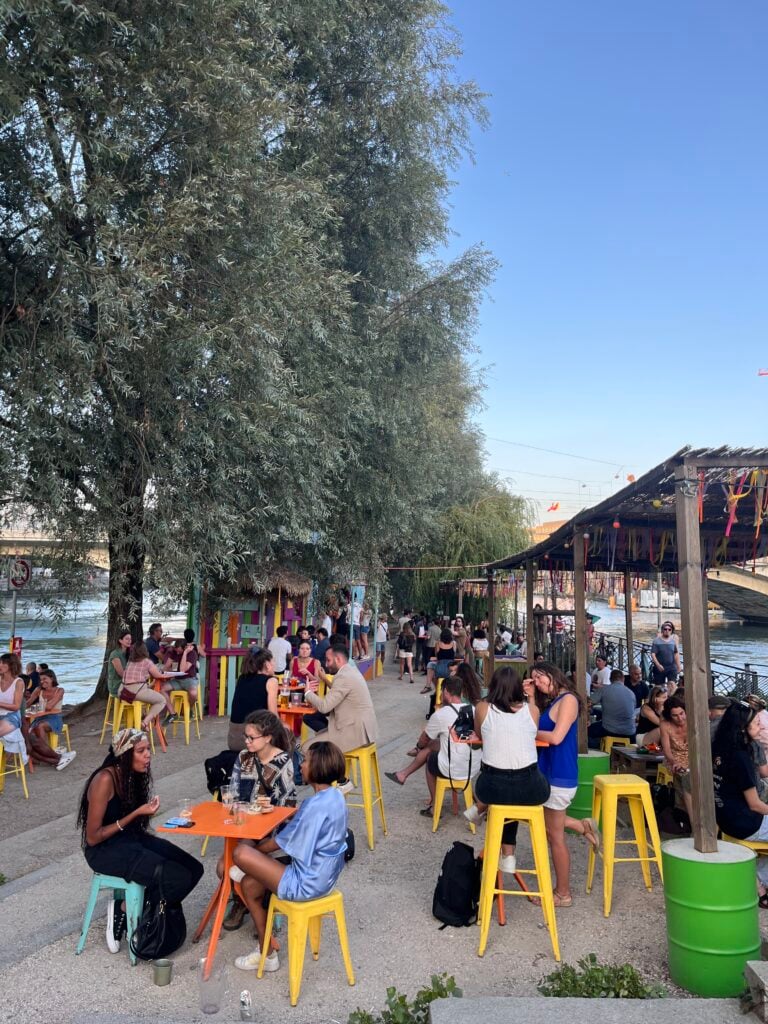 färglada stolar och bord och glada människor dricker drinkar på en udde med lummig grönska, vatten rinner längs med udden.