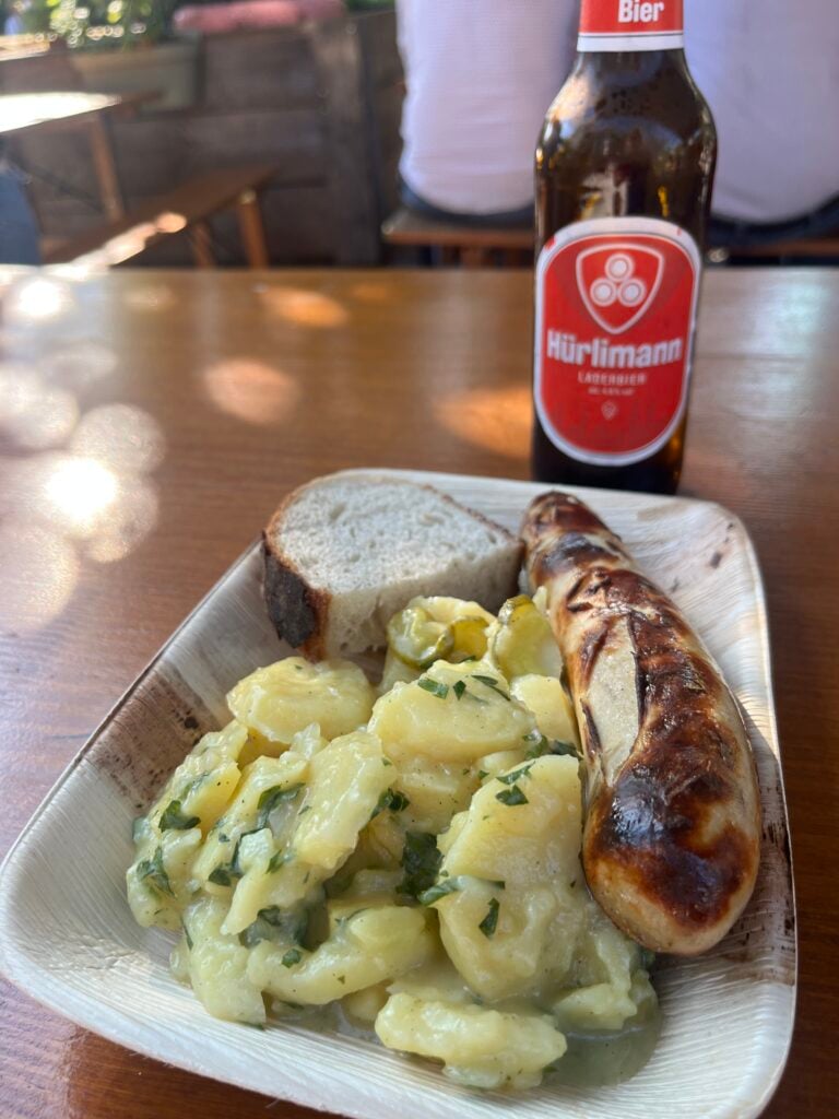 En korv med potatissallad på en engångstallrik och en flaska öl. 