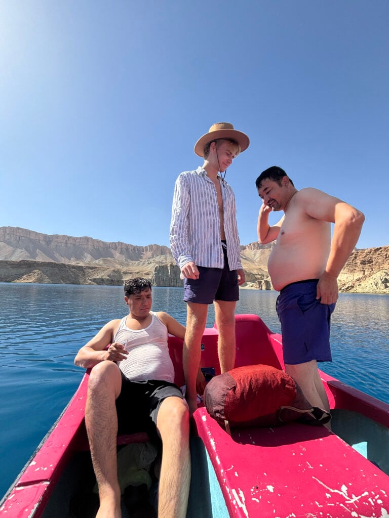 Tre killar i shorts står i en röd trampbåt på en blå sjö, omgiven av ökenberg.