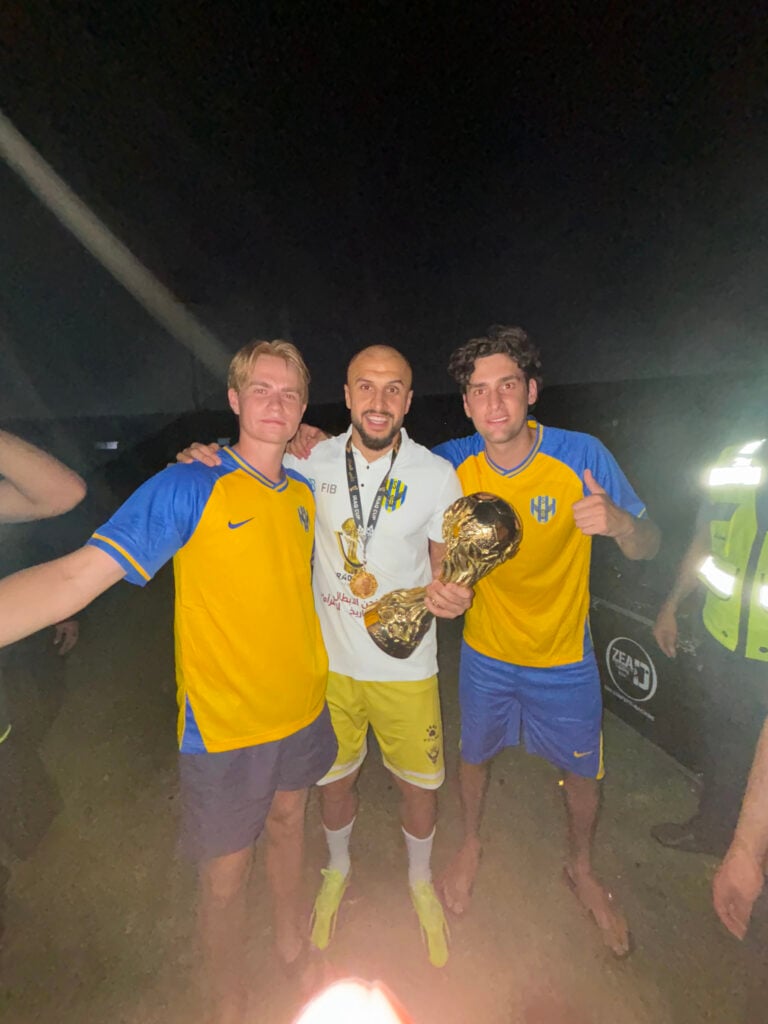 Två killar i gulblå fotbollskläder står med en vitklädd fotbollsspelare som håller i en fotbollspokal.