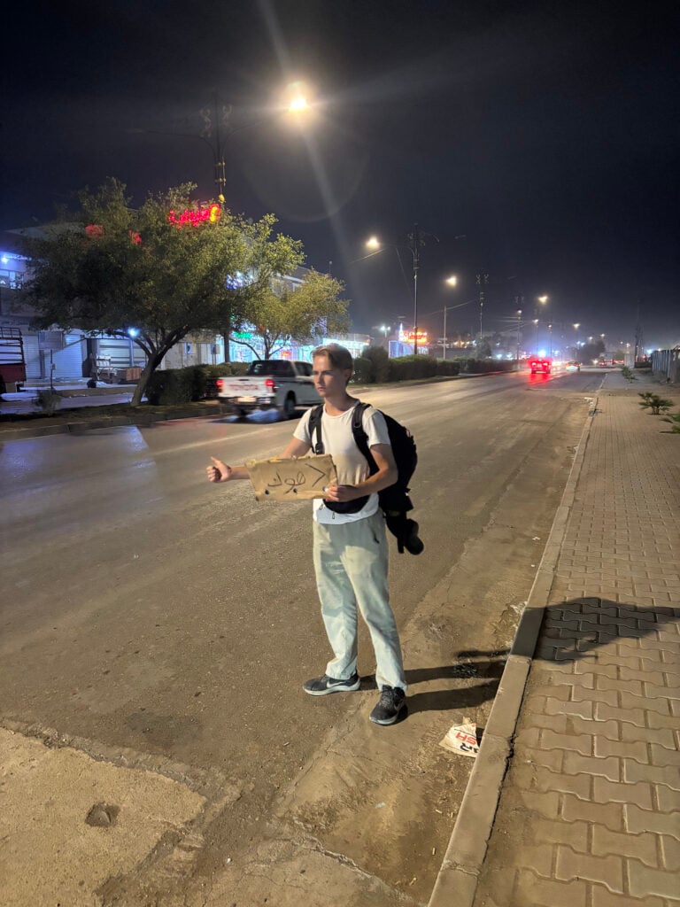 En person står i mörkret på en gata med en kartongbit med arabisk text. Han håller liftartummen upp. 