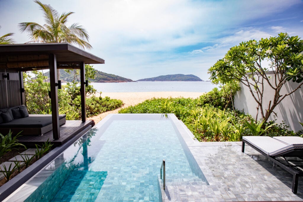 En turkos infinitypool syns framför en strand med tropisk grönska. 