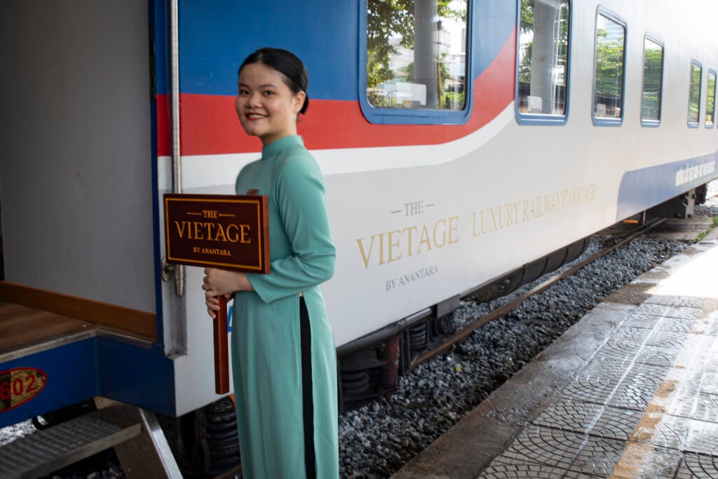 En kvinna i grönt med en skylt står utanför ett tåg i vitt, rött och blått. 