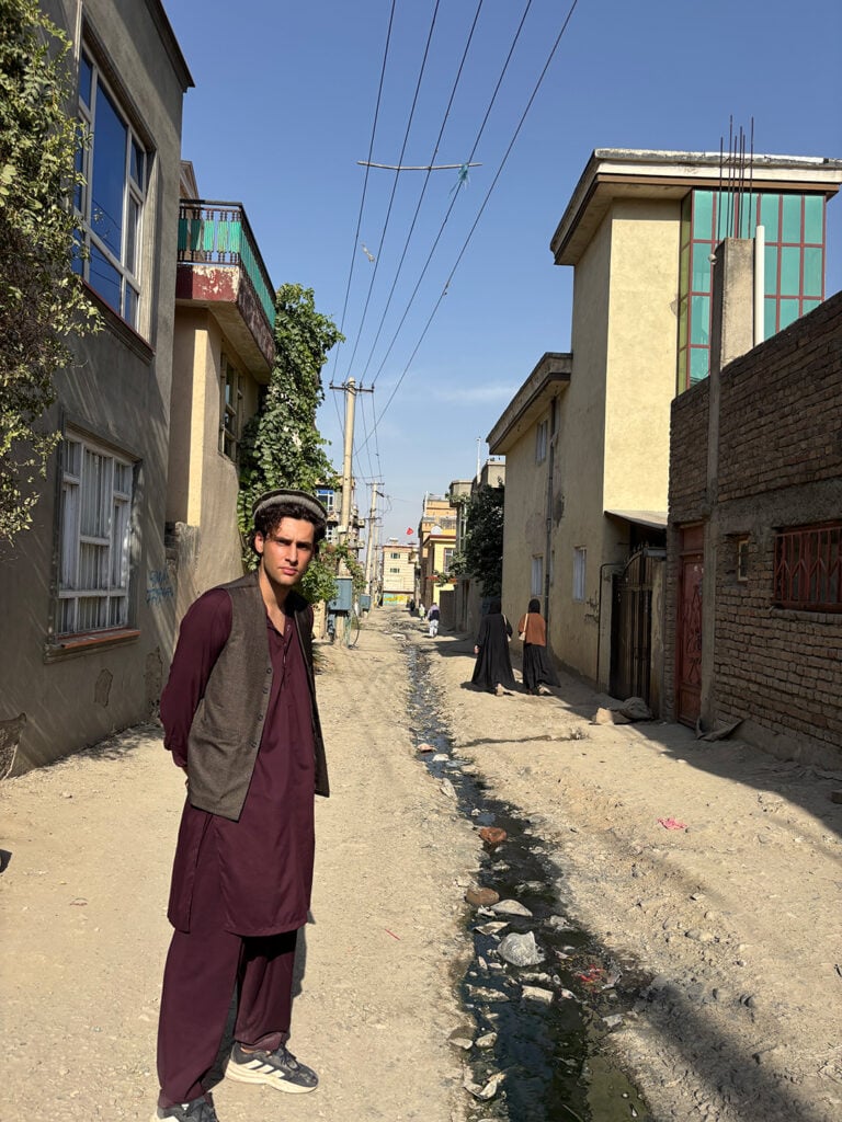 En kille i vinröda arabiska kläder och grå väst står i en dammig gränd där det rinner en blöt rännil i mitten. 