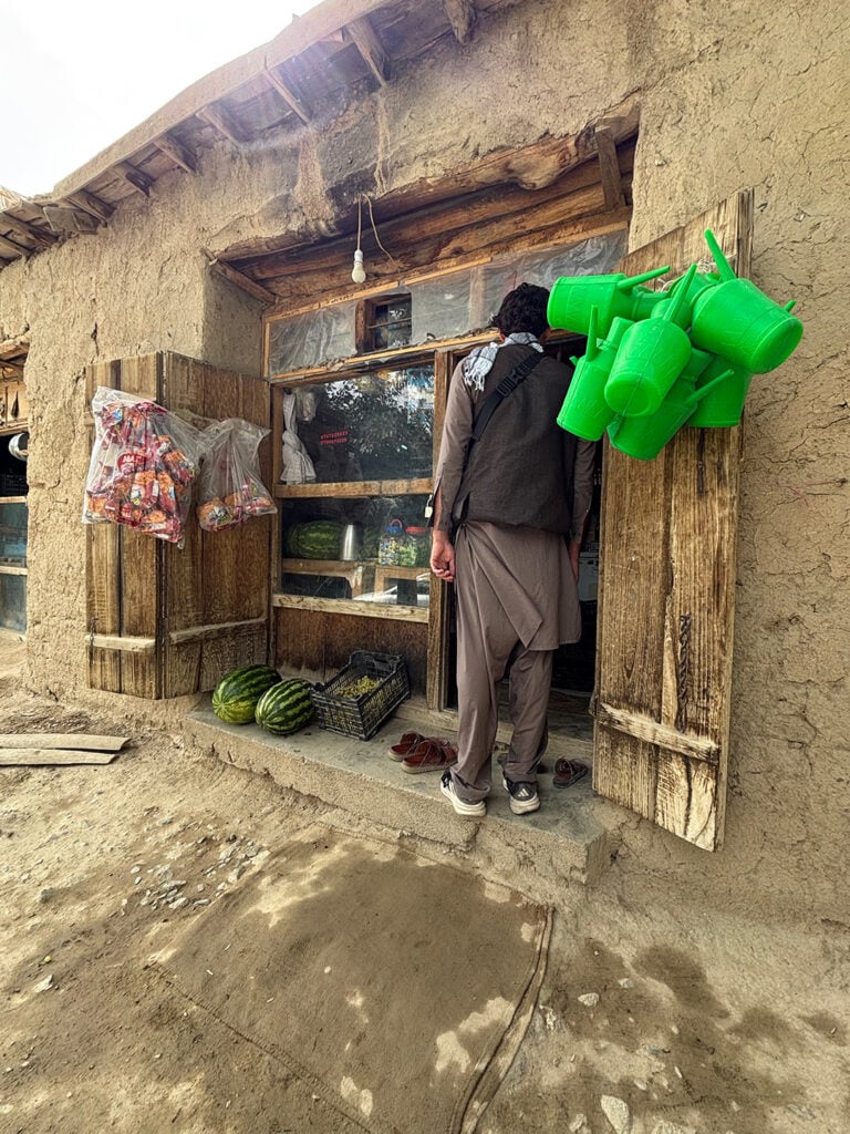 Ett hus med lerväggar avslöjar ett par grova trädörrar, glasfönster och ett knippe knallgröna vattenkannor som hänger utanför. 