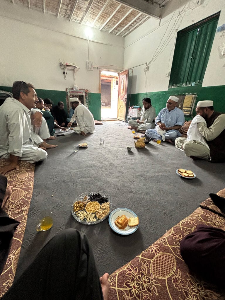 En rad äldre herrar i arabiska kläder och vita små hattar sitter på en matta i ett rum och äter. 