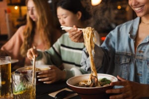 Woman lifting delicious steaming hot ramen noodles with chopsticks from bowl in restaurant with friends