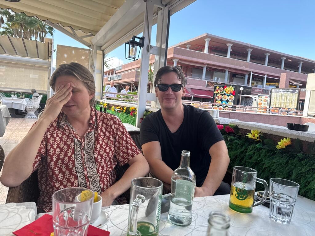 Två män sitter vid ett bord med flaskor och glas på en uteservering med betonghus bakom. 