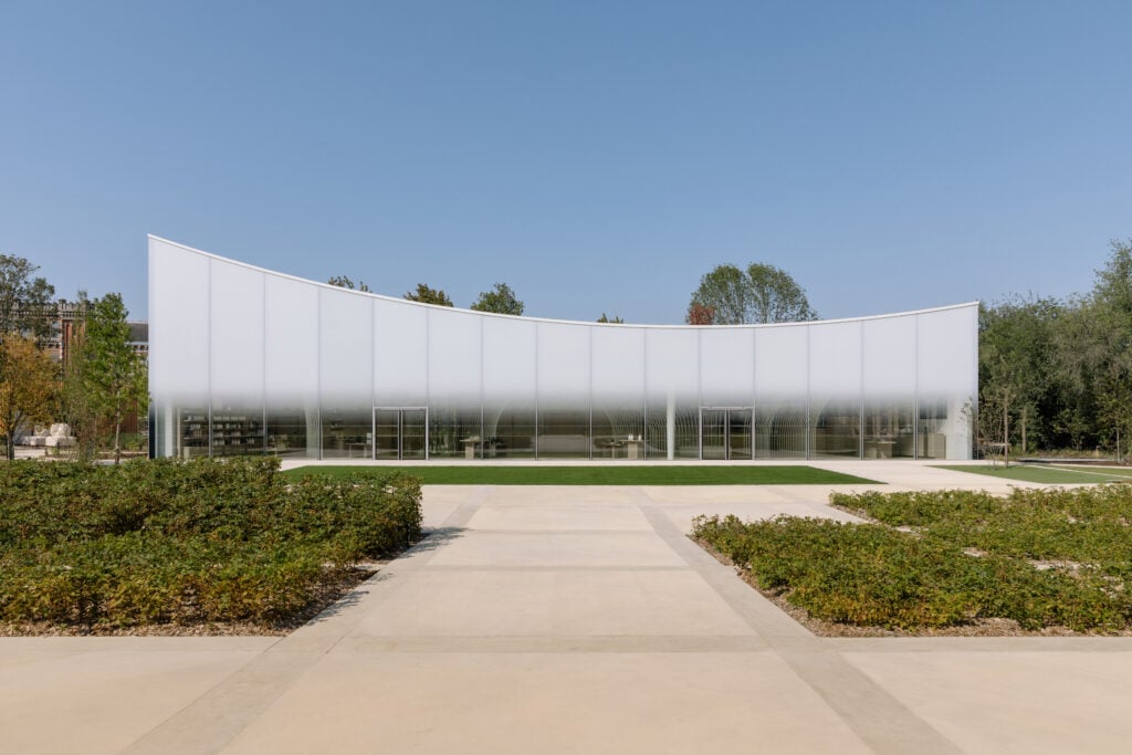En vit, välvd byggnad framför ett stenlagt torg med lite gröna planteringar. 