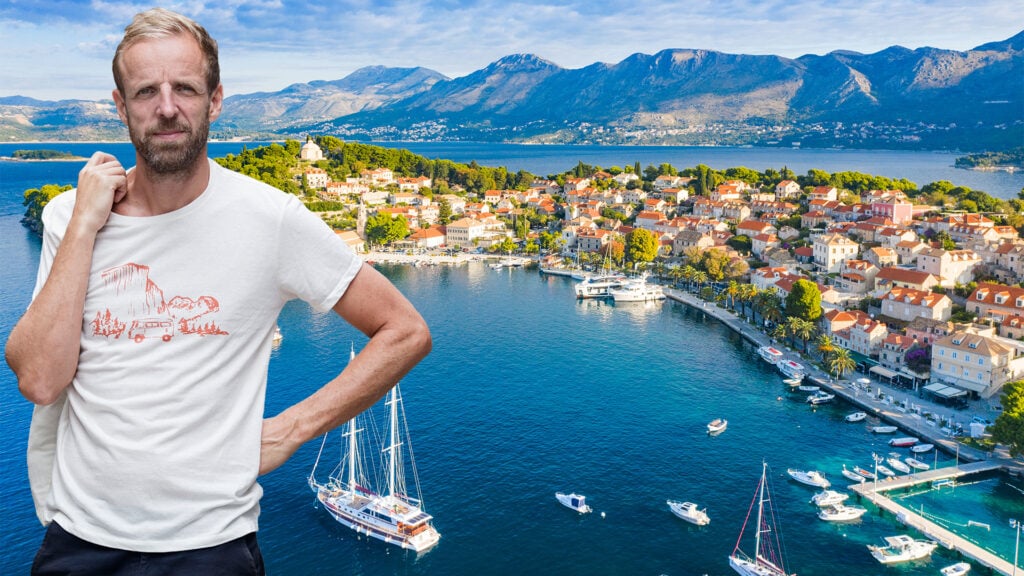 En drönarvy visar ett klarblått hav och en bebyggd, lummig halvö som sticker ut med ett dramatiskt bergslandskap på fastlandet bakom. Båtar och lyxyachter ligger för ankar. En man i vit t-shirt är inklippt framför bilden.