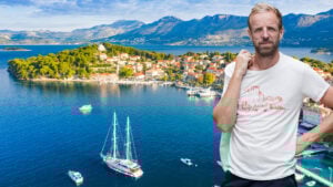 En drönarvy visar ett klarblått hav och en bebyggd, lummig halvö som sticker ut med ett dramatiskt bergslandskap på fastlandet bakom. Båtar och lyxyachter ligger för ankar. En man i vit t-shirt är inklippt framför bilden.