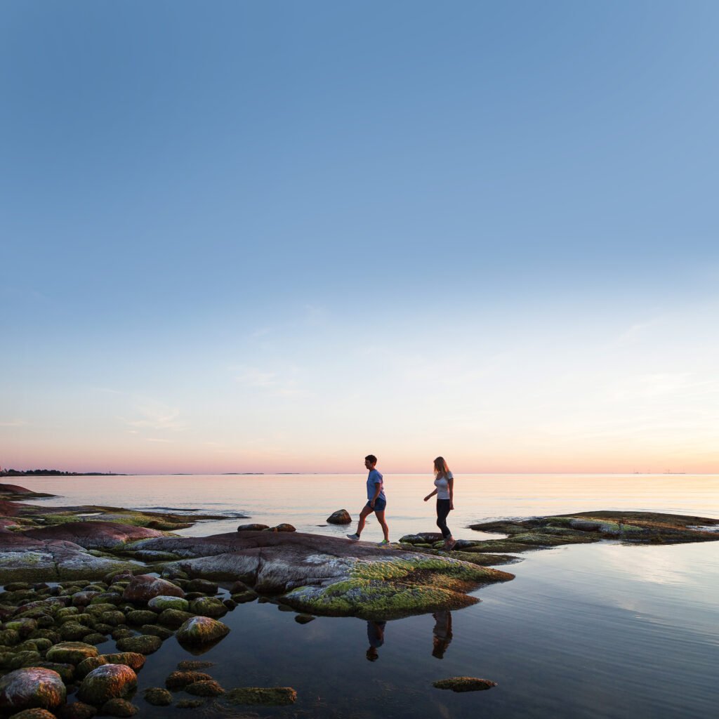 Två personer hoppar på klippor som sticker upp i ett spegelblankt hav i solnedgången.