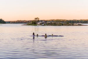 Barn och vuxna badar i havet bland klippor i solnedgången.