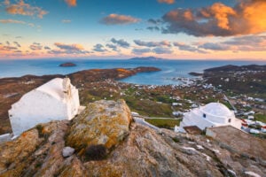 En vitkalkad gammal byggnad, högt upp på en klippa med utsikt över bergssluttningen, ett turkost hav i solnedgång.