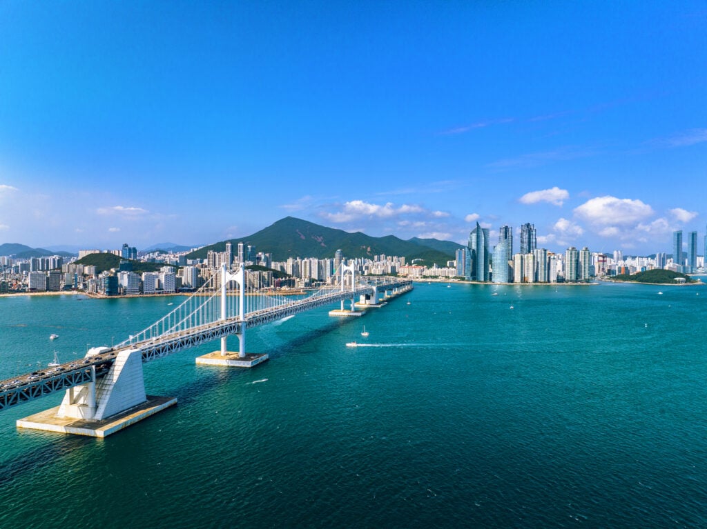 En vit hängbro går rakt ut i havet utanför en högteknologisk stad med skyskrapor, bergslandskap bakom.