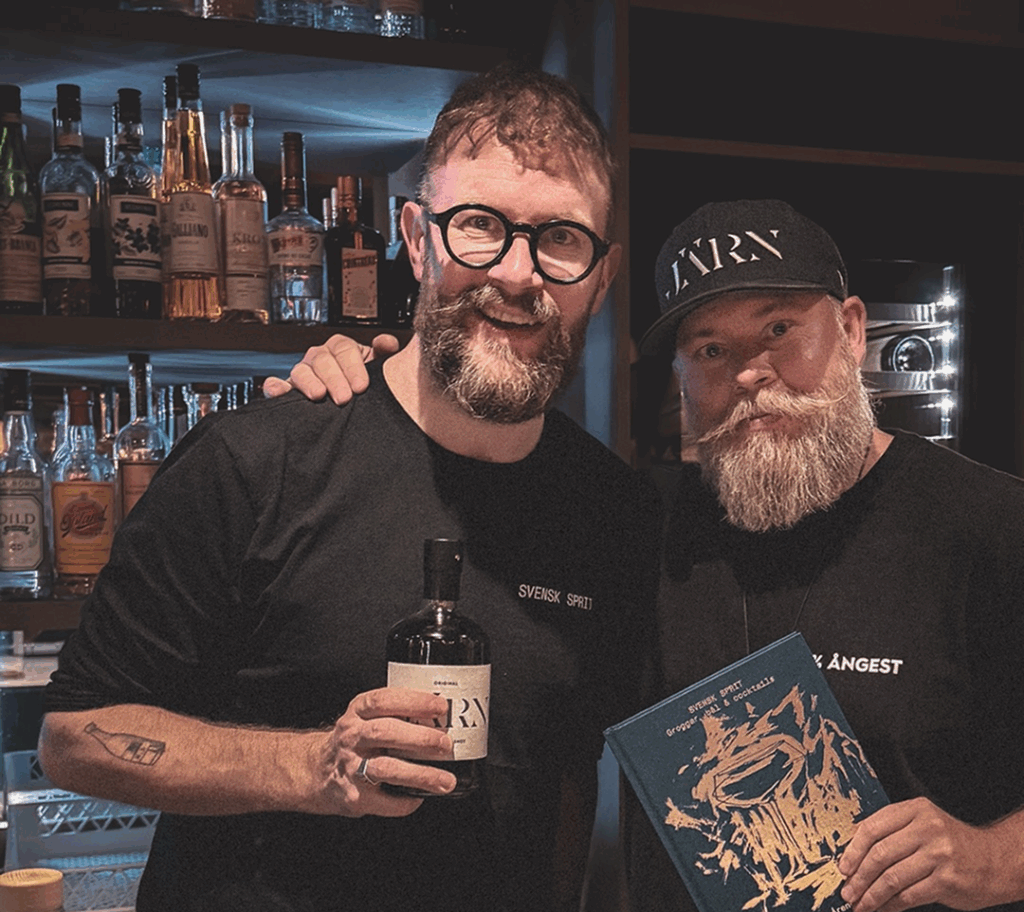 Två skäggiga män poserar och håller om varandra på en murrig bar. Den ena håller i en glasflaska, den andra i en bok. 