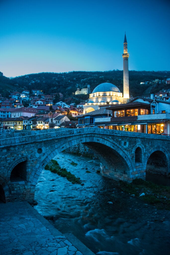 En stenbro över en flod framför en stad med ett lummigt berg, i kvällsljus.