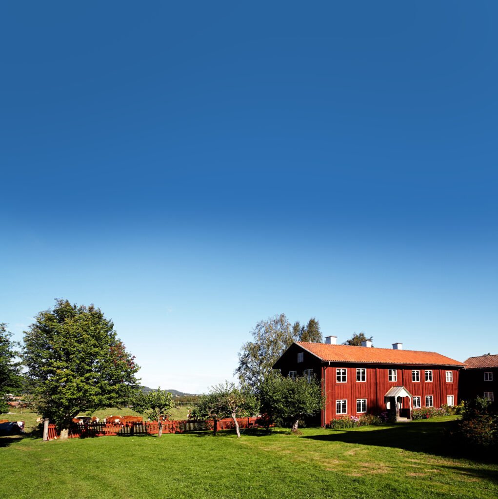 En stor, röd gård, grön gräsmatta och lummiga träd, rött staket, blå himmel.