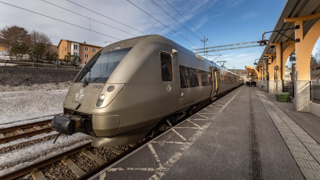 Ett grått tåg står på en perrong, blå himmel. 