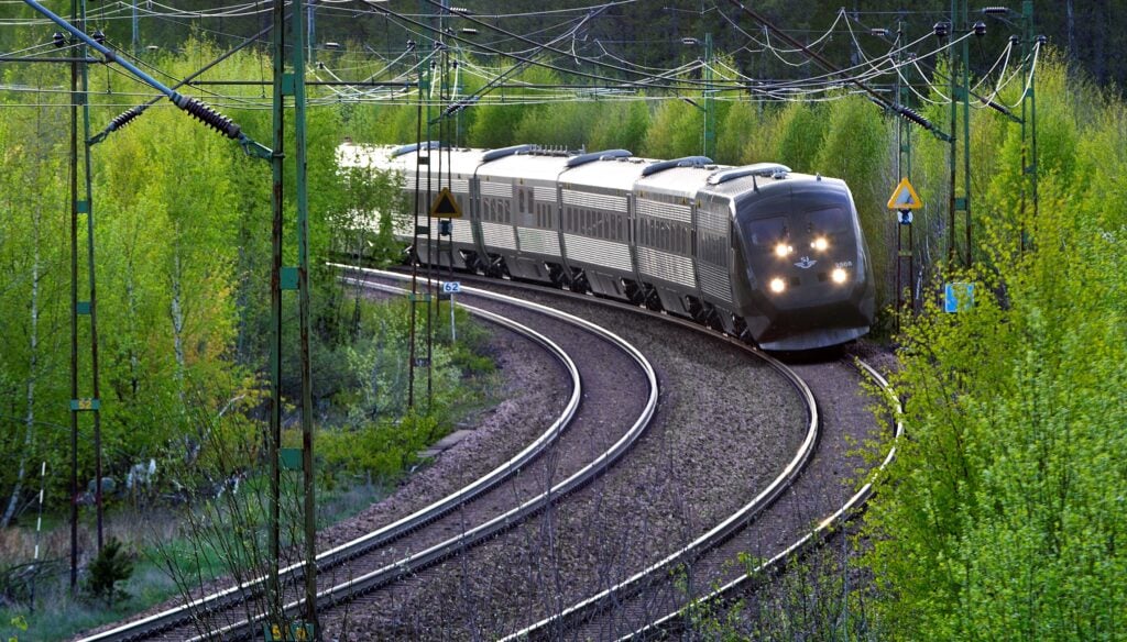 Ett tåg susar fram på spår mellan gröna träd. 