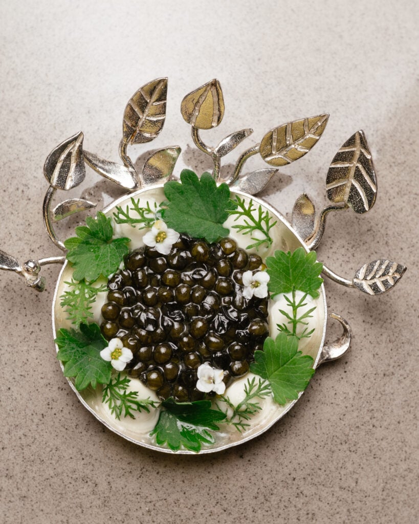 En vit tallrik, med silverfärgade skulpterade löv, tallriken är fylld av mörkgröna oliver, pyntad med grönt och vita blommor.