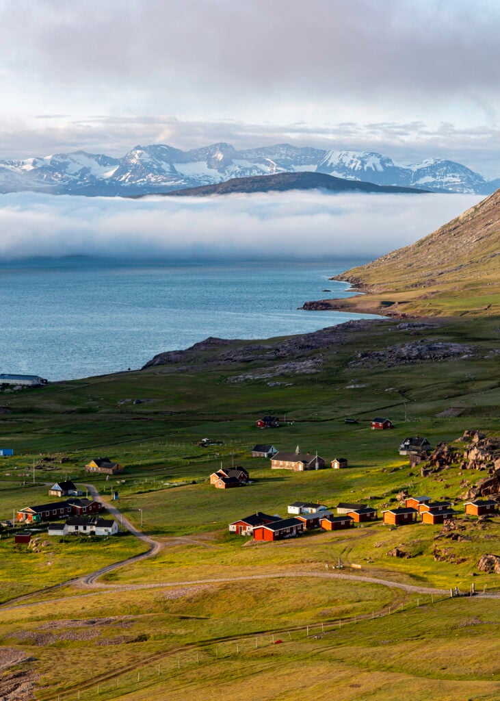 En ödslig bygd i en sluttning, ner mot havet. Dramatiska snötäckta berg i bakgrunden.