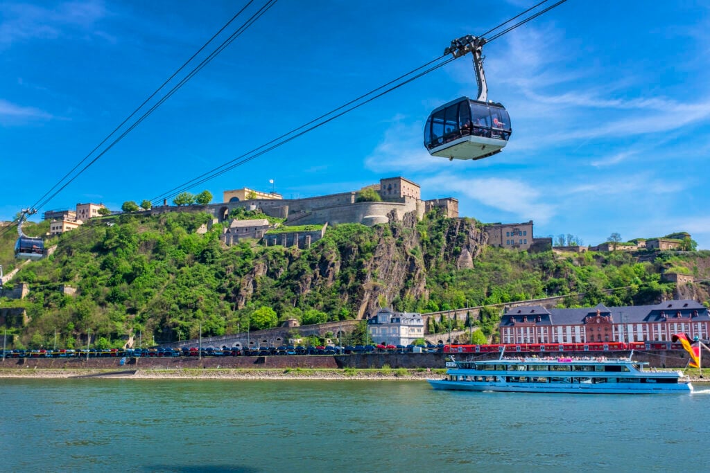 En flod med ett fartyg längs med stranden, ett rött tåg. En dramatisk klippa bär upp ett fort. En linbana går över himlen. 