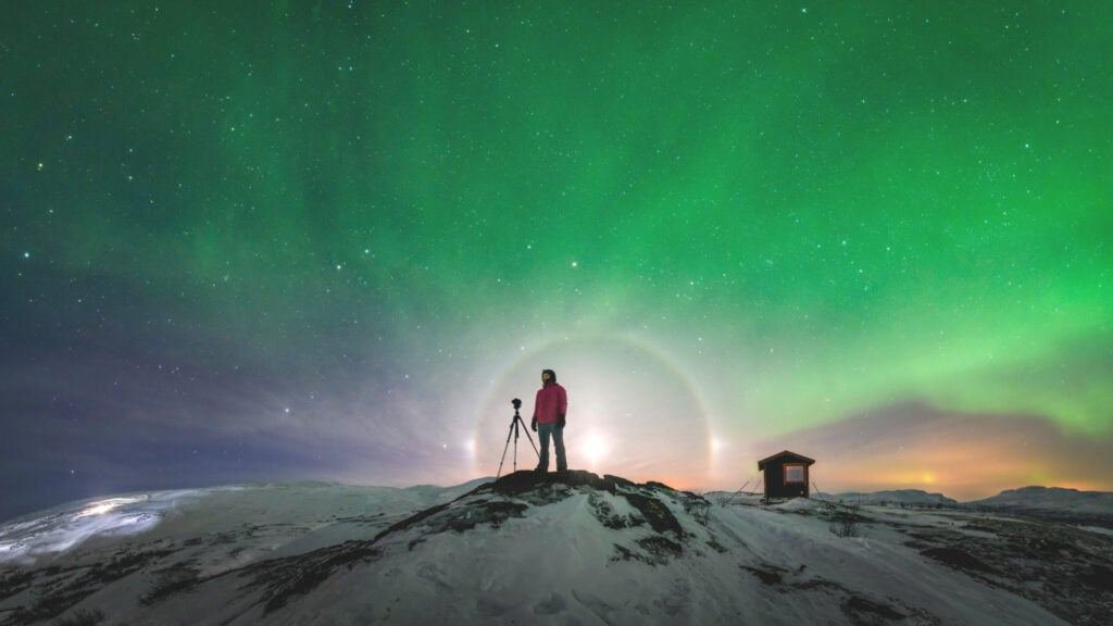 En person står framför en ljusbåge på en snötäckt klippa under en norrskensgrön himmel.