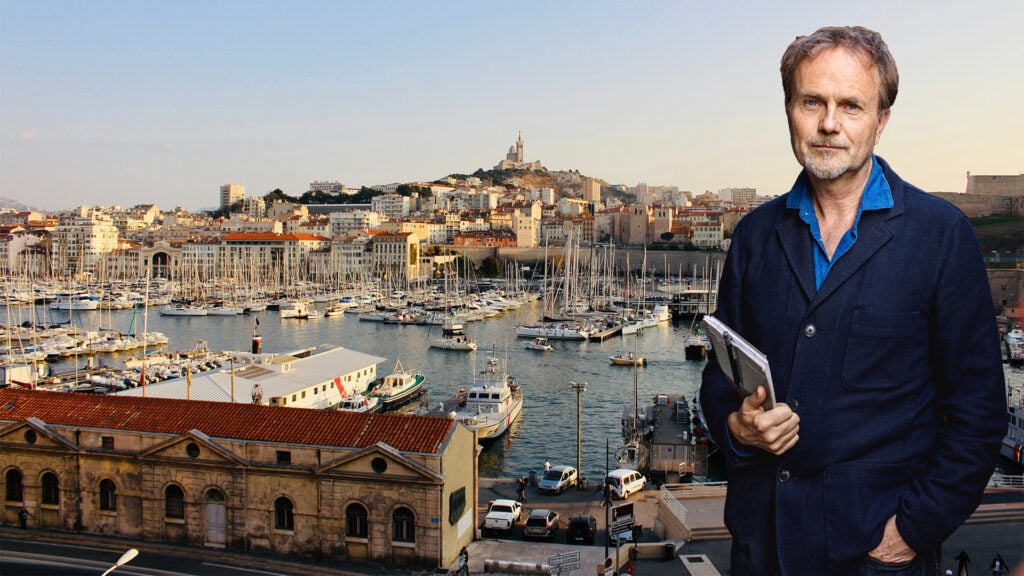 Marseille från en hög vy, vatten, båtar, en kulle med mycket byggnader. En inklippt man poserar bredvid bilden.