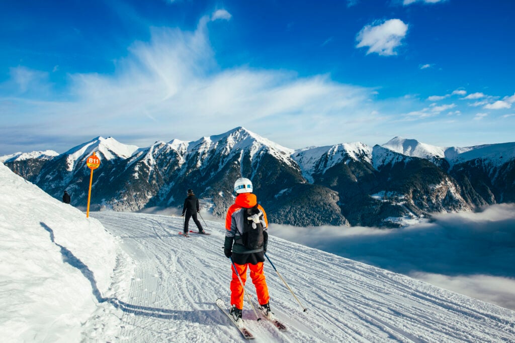 En skidåkare i orange och en i svart glider fram framför Alperna. 