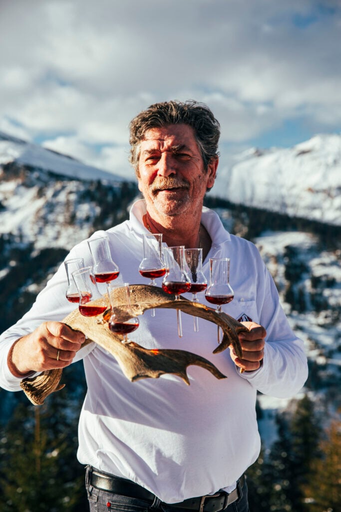En man i vit tröja håller upp spritfyllda glas i ett hjorthorn, framför Alperna. 