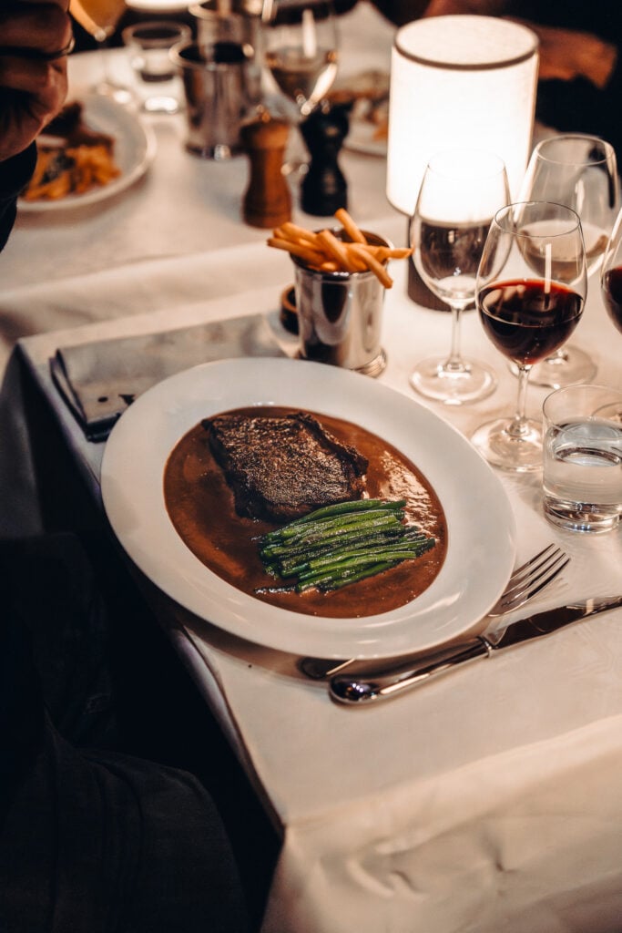 En tallrik med en biff och gröna grönsaker, på en spegel av sås. Bredvid står en burk med pommes. Bordet är dukat med vita dukar och vin.