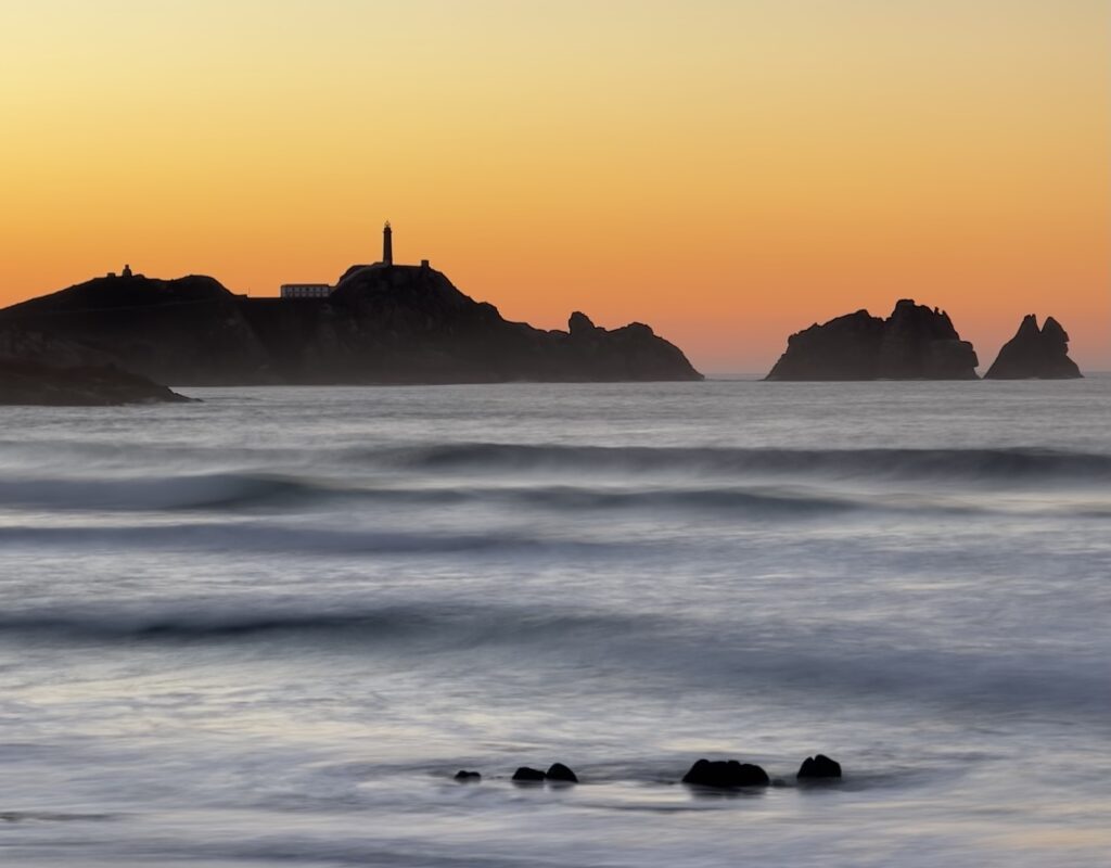 En fyr på en hög klippa framför en orange solnedgångshimmel, ett blått hav framför. 
