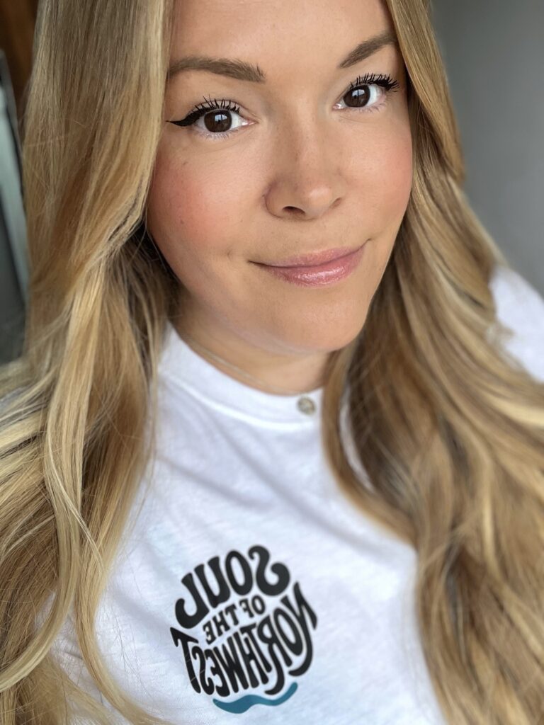 En kvinna med blont hår och vit t-shirt ler mot kameran. 