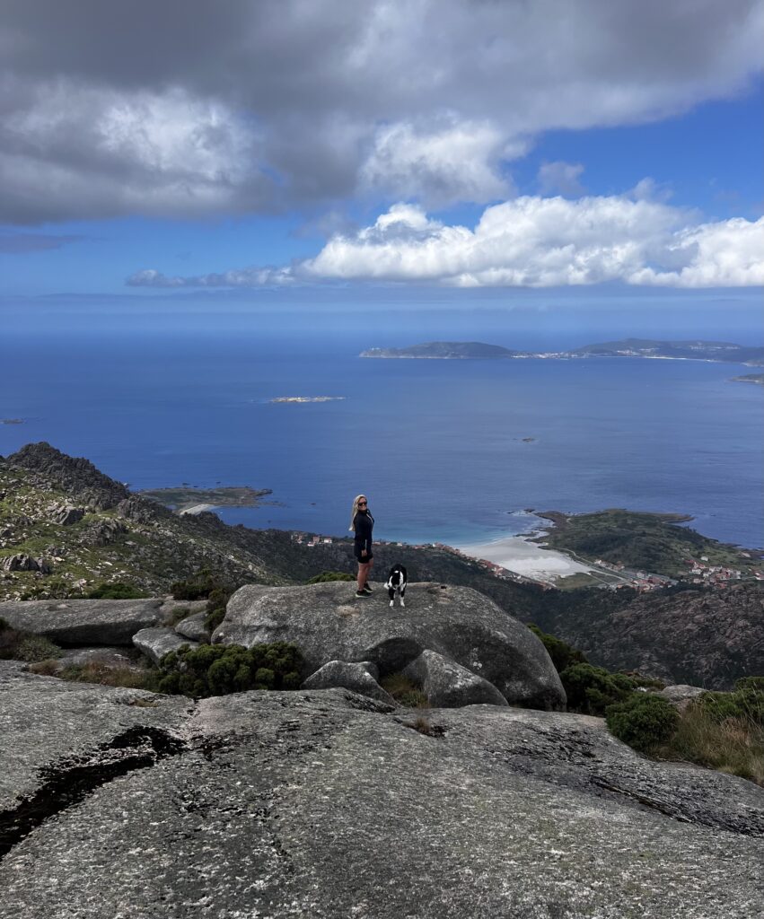 En kvinna och hennes hund står på en bergstopp och blickar ut över havet.