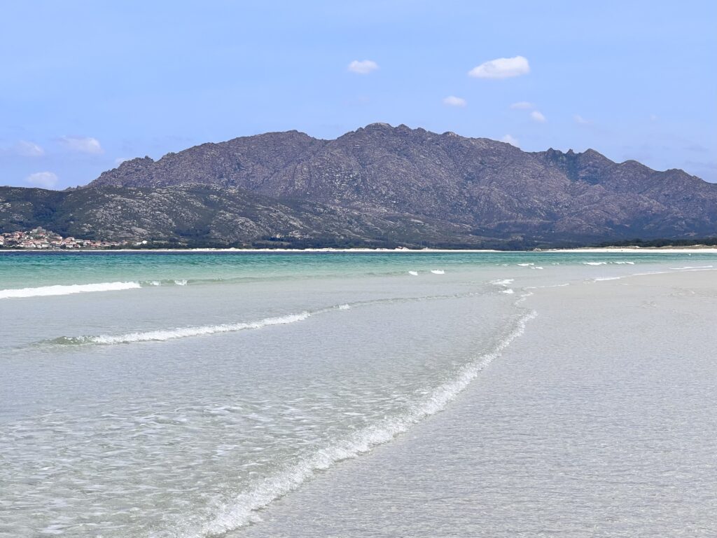 En vit sandstrand med kristallklart vatten, höga kullar bakom. 