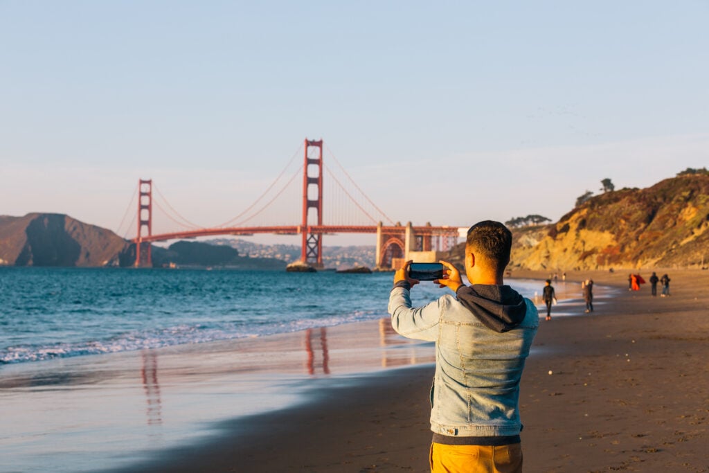 En man står på en strand och fotar Golden Gate.