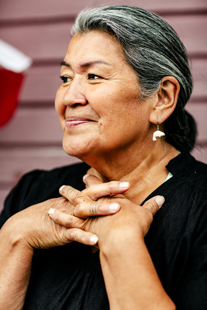 En kvinna med gråsvart hår och små vita sälar i öronen ler och knäpper händerna över bröstet.